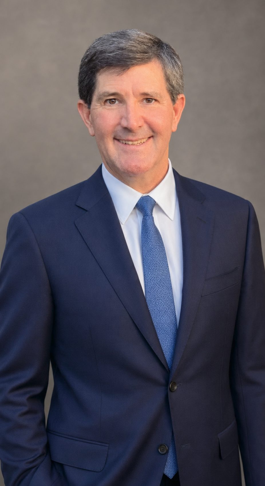 Rosser Newton standing in a navy suit and blue tie against a plain grey studio backdrop